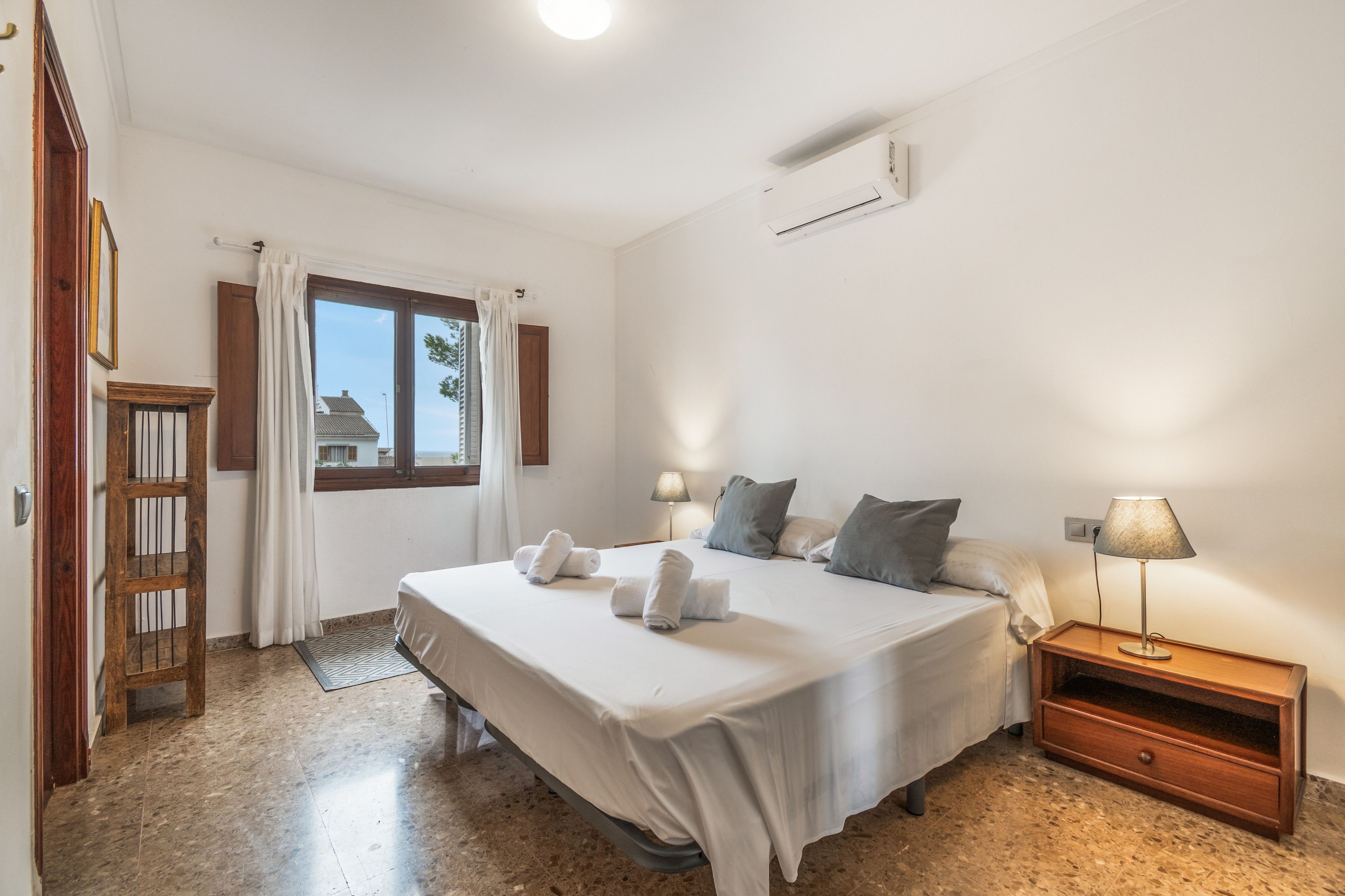 Ein Hotelzimmer mit einem Bett, zwei Nachttischlampen, einem Nachttisch, einem Holzschrank, einem Fenster mit Vorhängen und einer Klimaanlage.