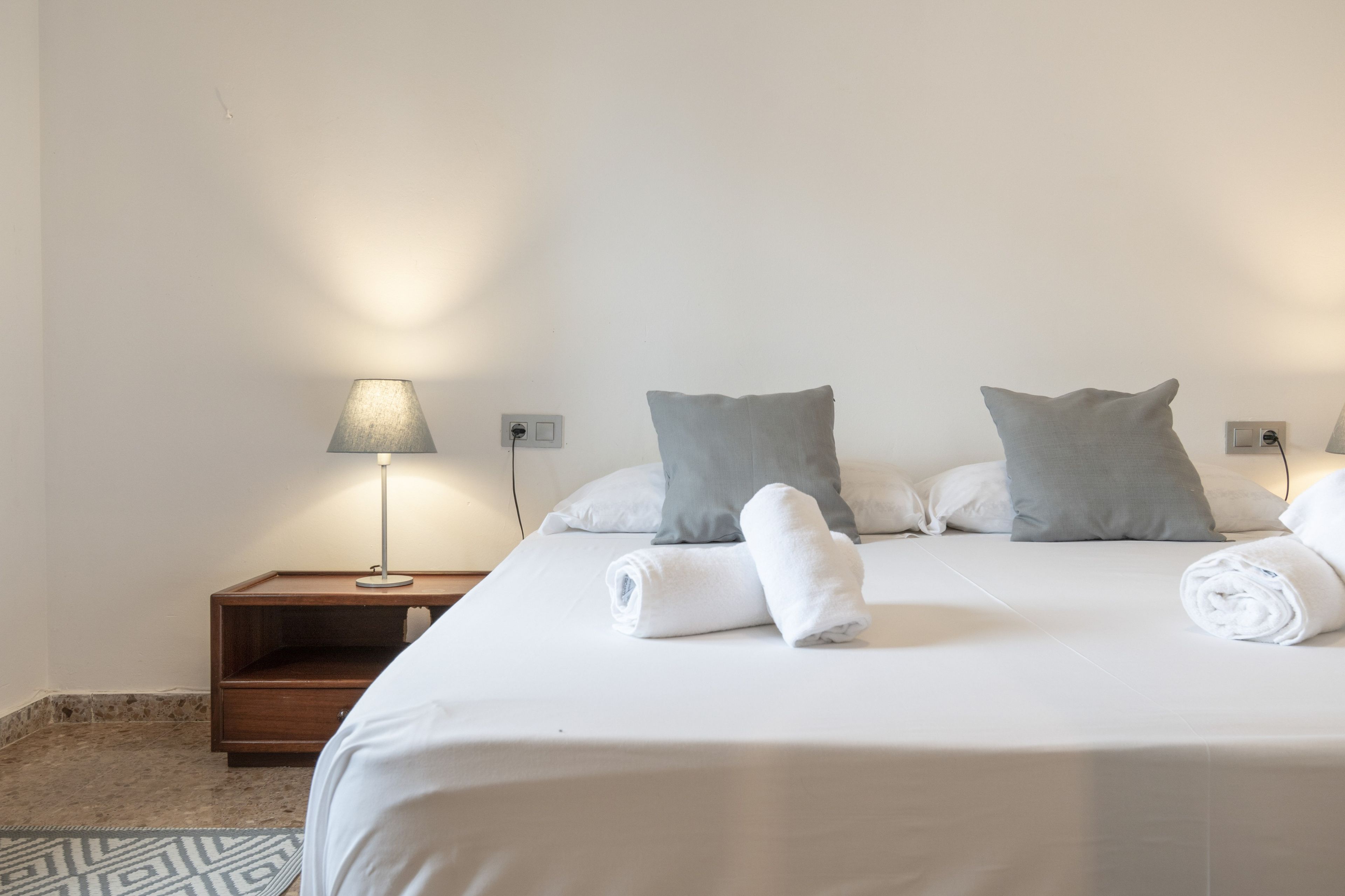 A neatly made bed with white linens and rolled towels, a bedside lamp, and a wooden nightstand.