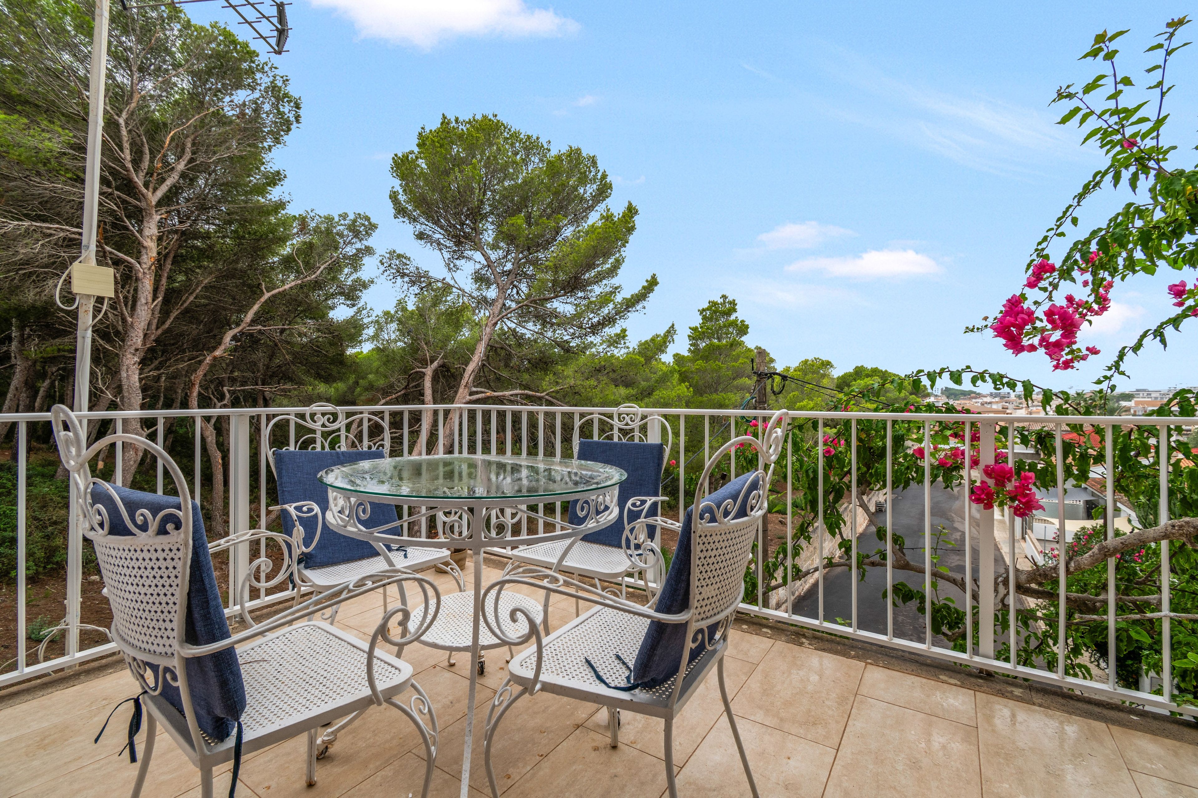 An outdoor patio with a round glass table and white metal chairs, surrounded by a metal railing.