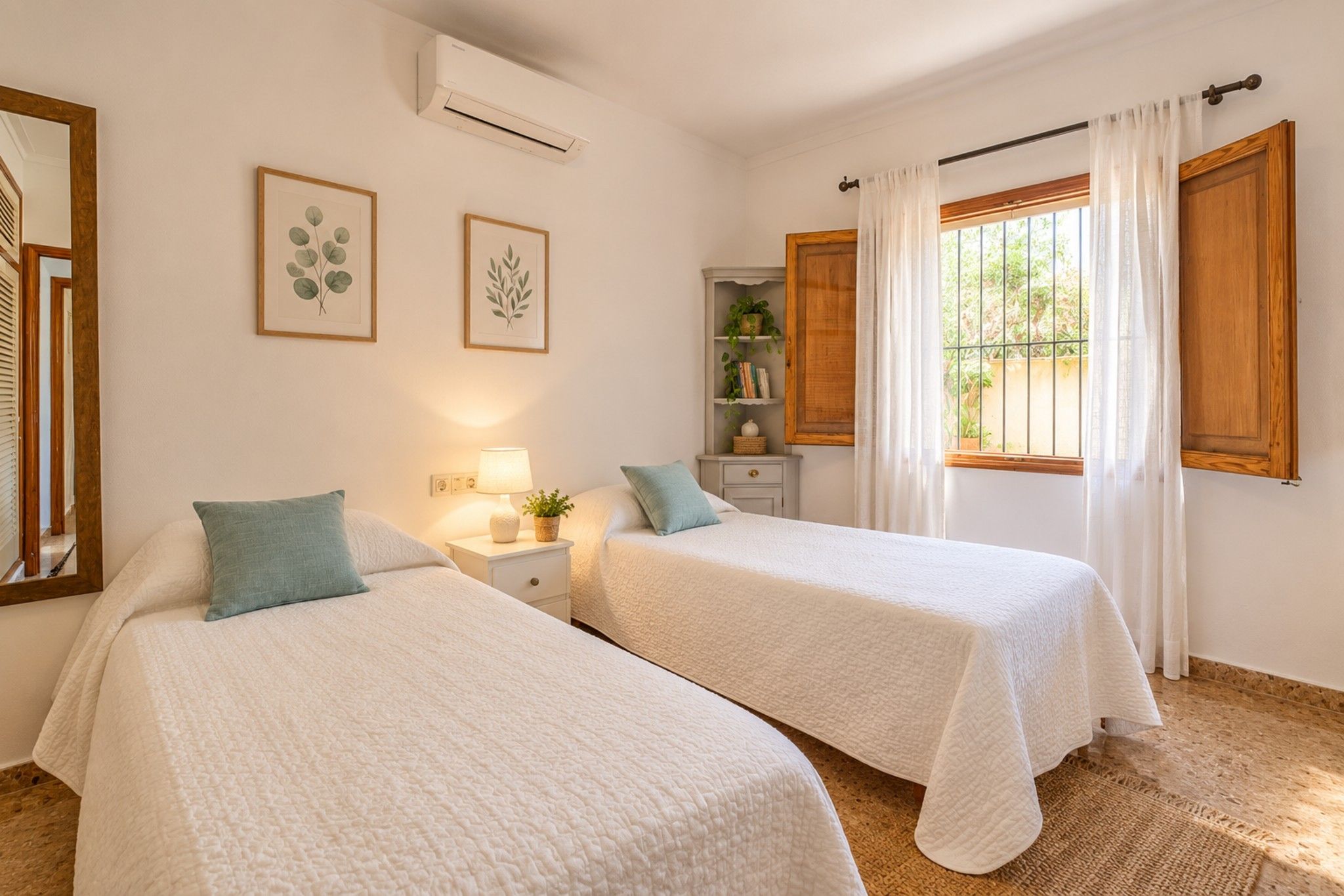 Quarto de hotel com duas camas, uma mesa de cabeceira com abajur, um pequeno armário com livros e uma planta, e uma janela com cortinas.