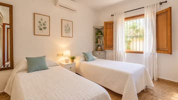 Quarto de hotel com duas camas, uma mesa de cabeceira com abajur, um pequeno armário com livros e uma planta, e uma janela com cortinas.