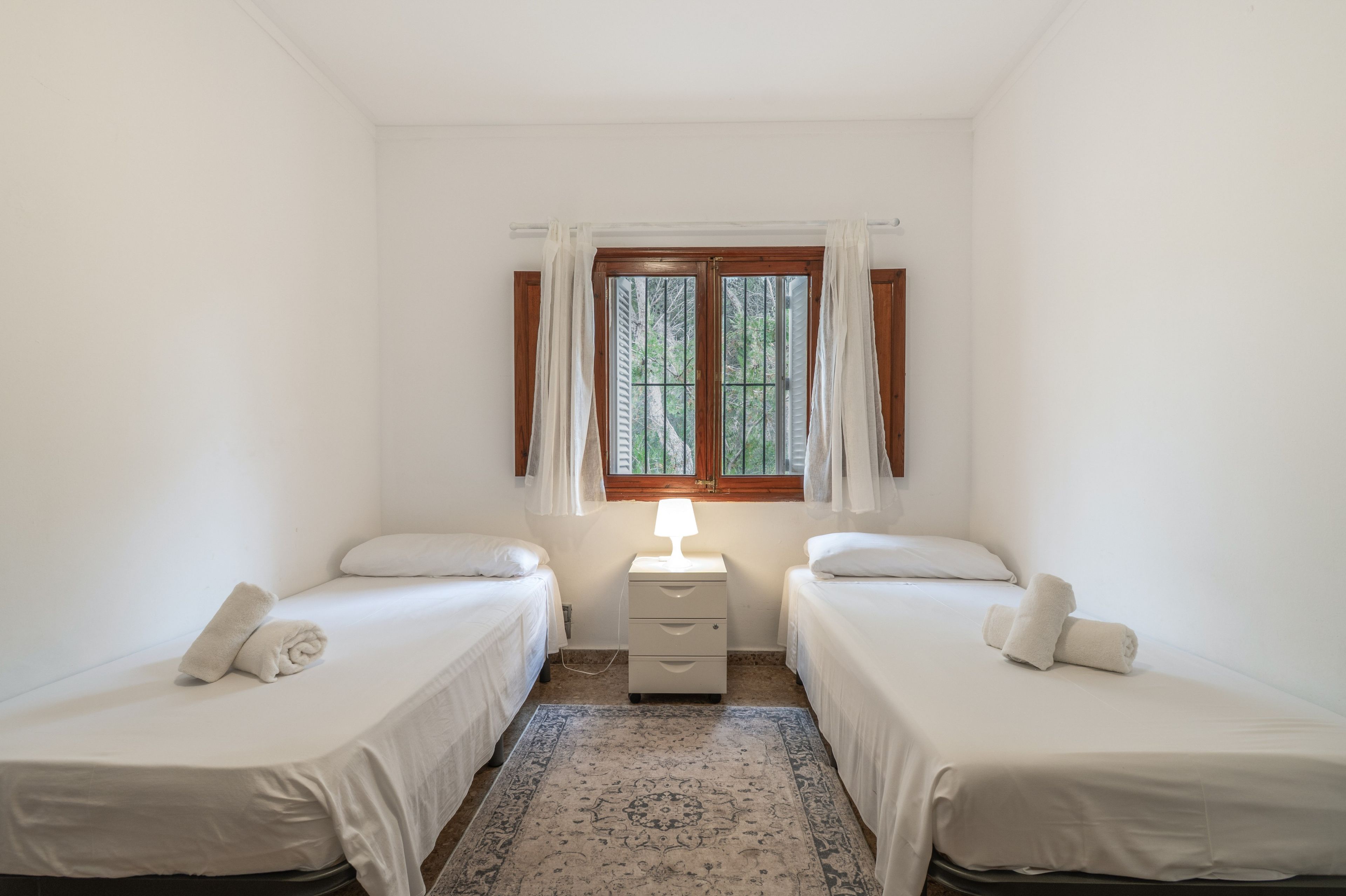 A room with two single beds, a nightstand with a lamp, a window with curtains, and a patterned rug.
