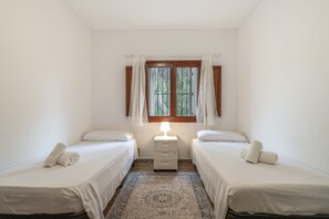A room with two single beds, a nightstand with a lamp, a window with curtains, and a patterned rug.