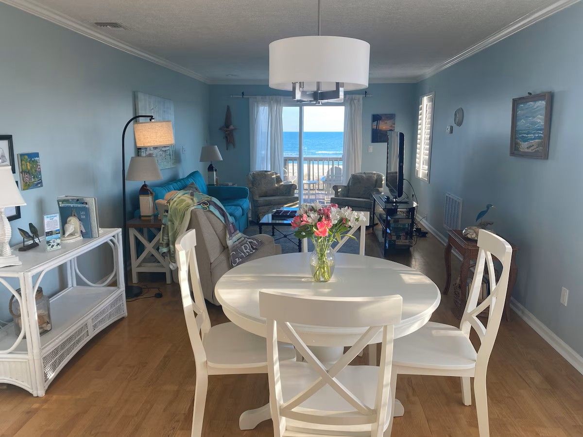 A room with a round dining table, white chairs, a white console table, and a view of the ocean through large windows.