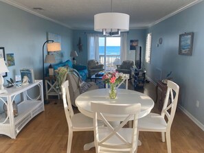 A room with a round dining table, white chairs, a white console table, and a view of the ocean through large windows.