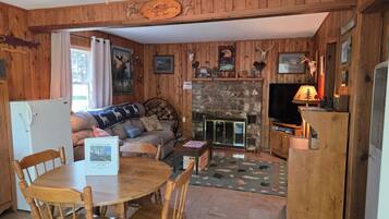 A cabin interior with a stone fireplace, wooden walls, a television, and a dining area with a table and chairs.