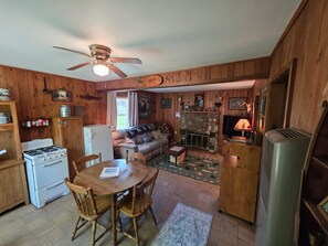 A cabin interior with a dining table, chairs, a sofa, a fireplace, a ceiling fan, and wooden walls.