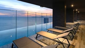 Un espace piscine intérieur moderne, doté de murs en verre, de chaises longues et offrant une vue sur la mer.