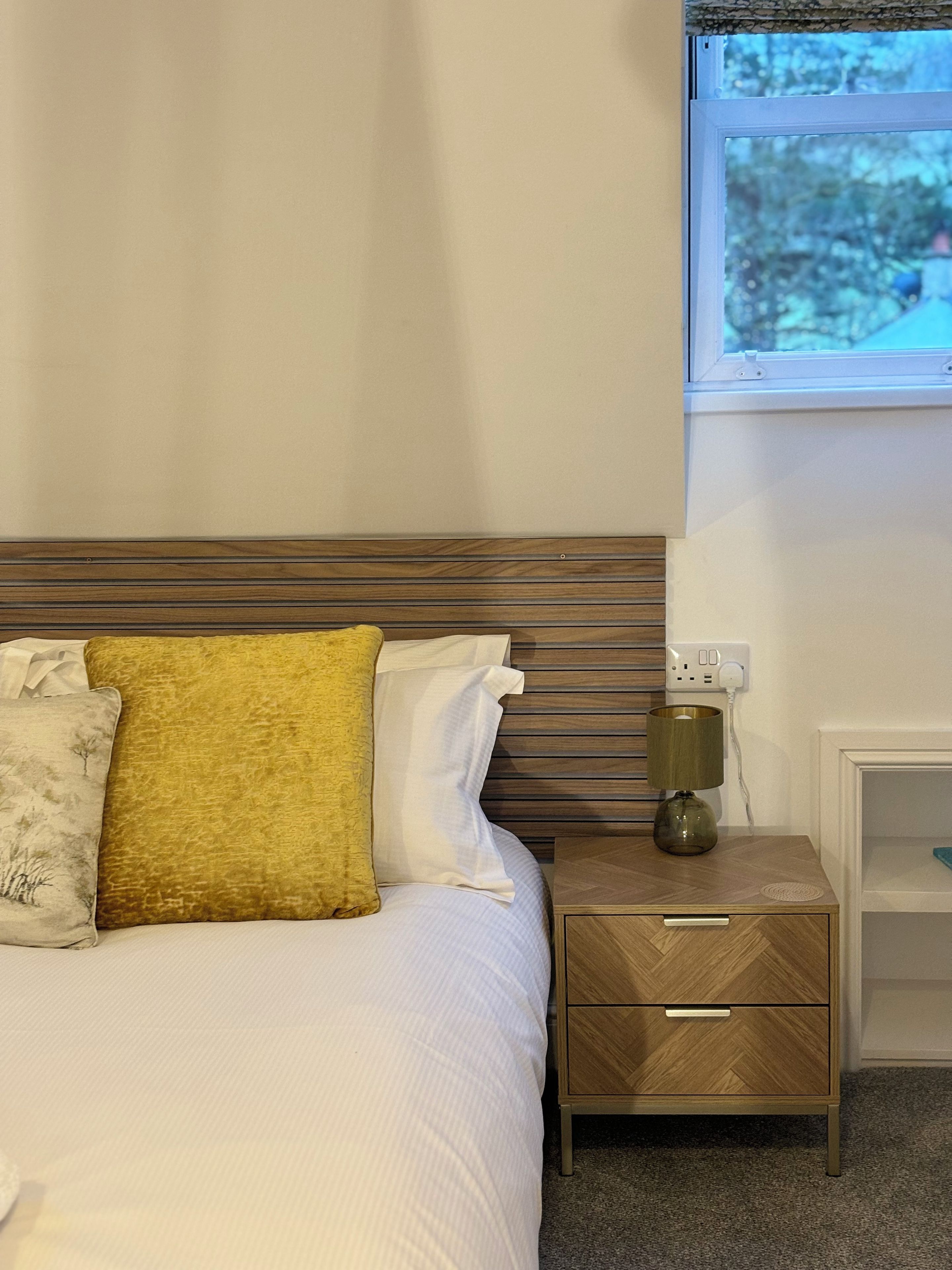Una cama bien hecha con cabecera de madera, una mesita de noche con lámpara y un enchufe en la pared.