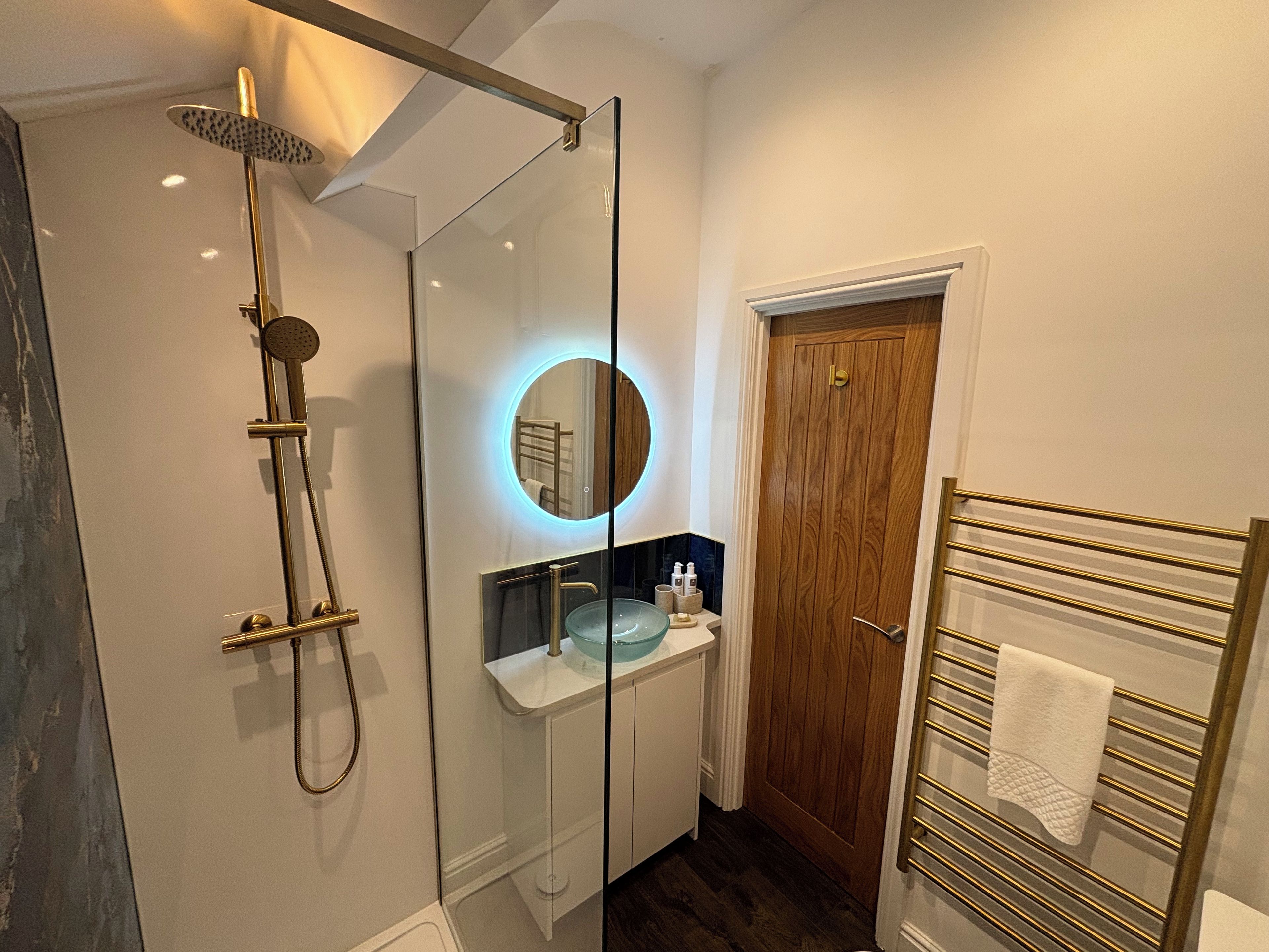 A modern bathroom with a glass-enclosed shower, a round mirror with a blue light, a wooden door, and a towel rack.
