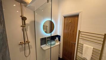 A modern bathroom with a glass-enclosed shower, a round mirror with a blue light, a wooden door, and a towel rack.