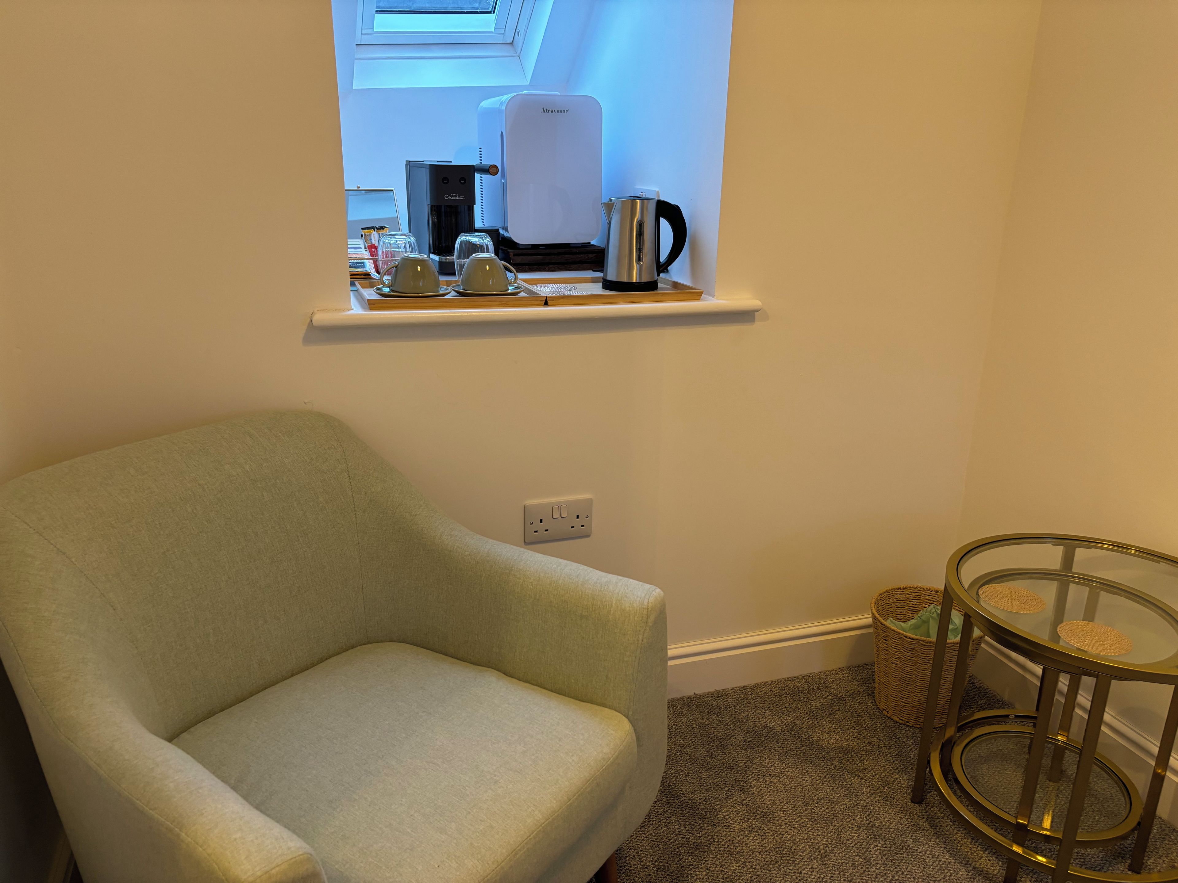 A room with a light green armchair, a coffee maker, and a small table with a glass top.