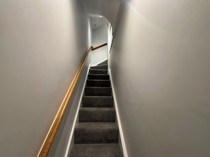 A staircase with carpeted steps and a wooden handrail, leading upwards in a narrow hallway.