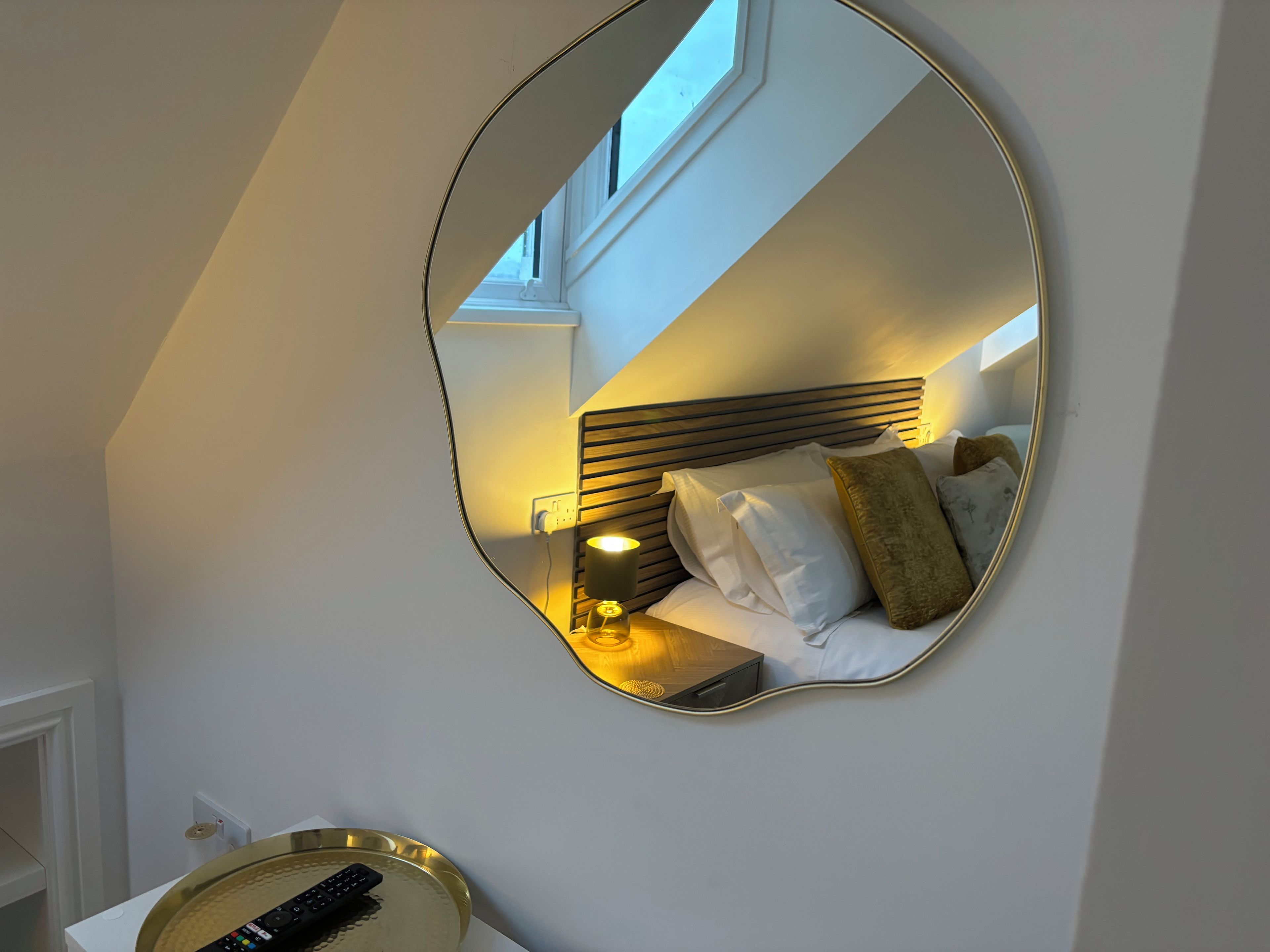 Ein Schlafzimmer mit einem Bett, einem Nachttisch, einer Lampe und einem an der Wand befestigten Spiegel.