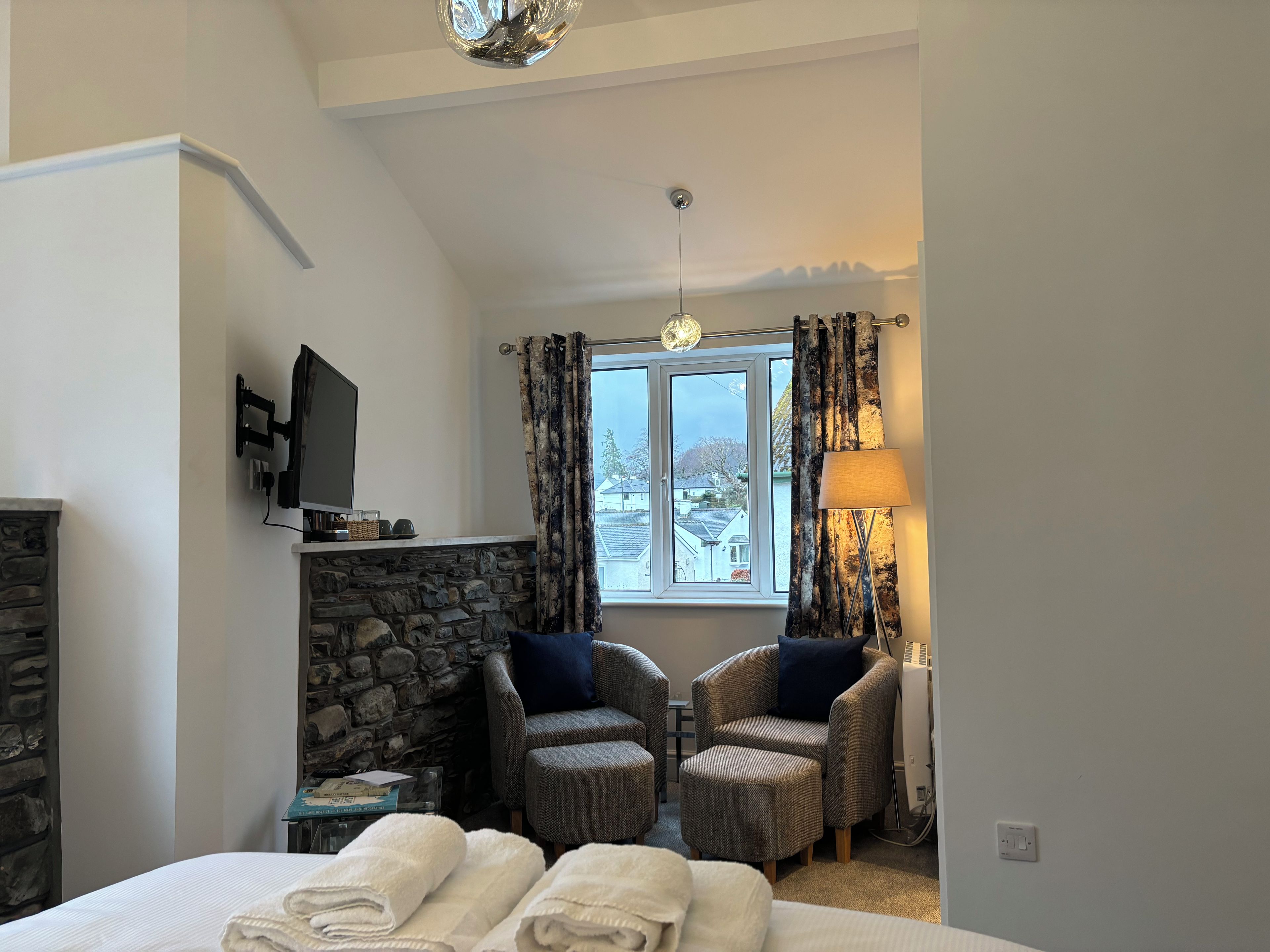Quarto de hotel com lareira de pedra, televisão, duas poltronas, janela com cortinas e cama com toalhas.