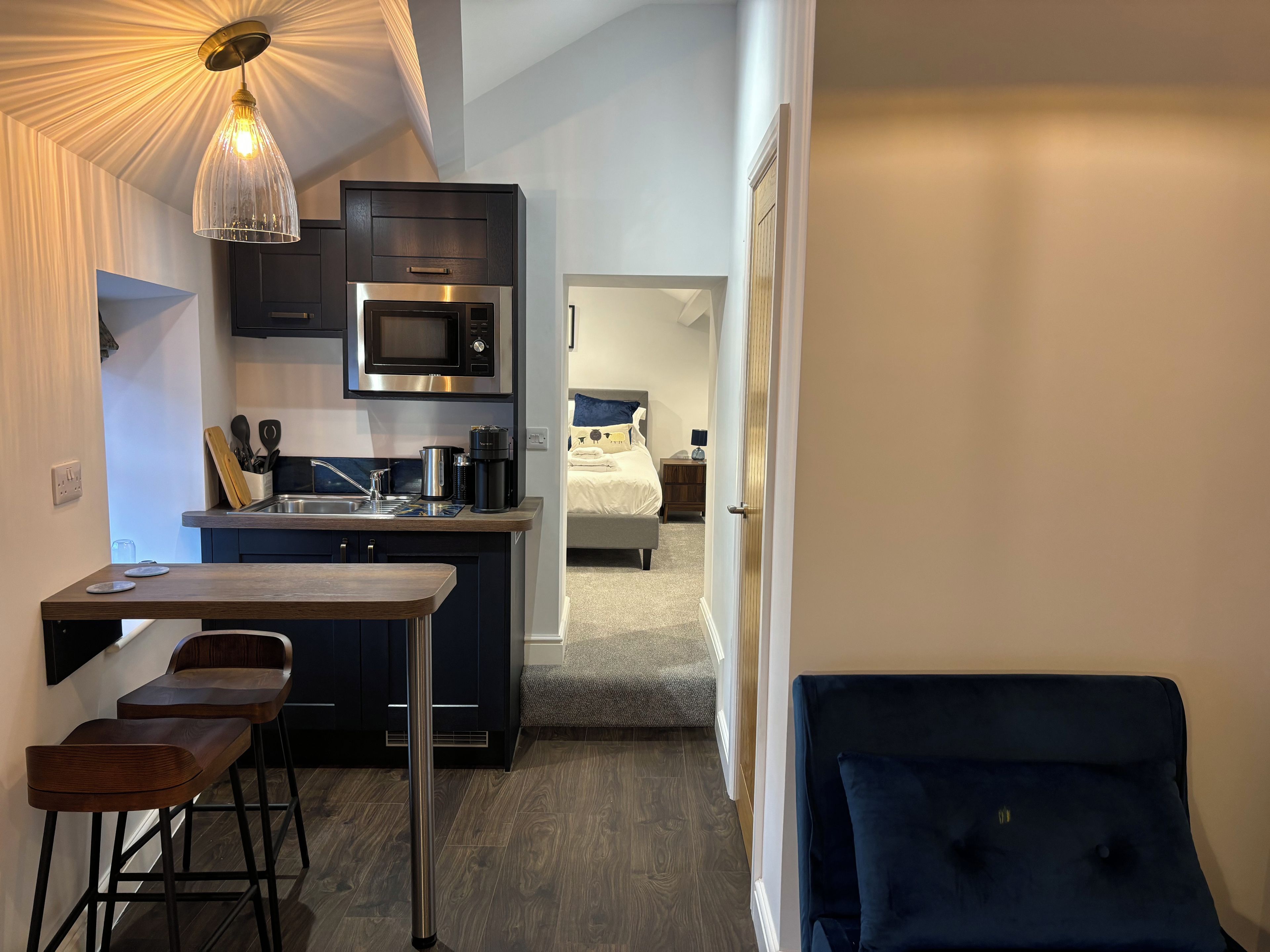 Cozinha compacta com micro-ondas, cafeteira e banquetas. Uma cama com travesseiro azul é visível no quarto adjacente.