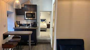 Cozinha compacta com micro-ondas, cafeteira e banquetas. Uma cama com travesseiro azul é visível no quarto adjacente.