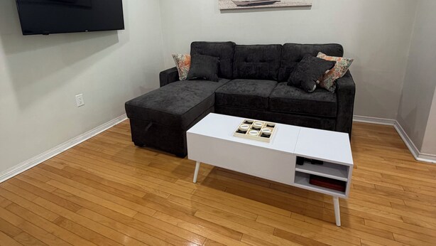 Un salon avec un téléviseur à écran plat fixé au mur, un canapé d’angle noir avec des coussins, une table basse blanche avec un plateau, et un tableau représentant une scène de plage.