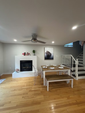 Amplia sala de estar con chimenea, una mesa de comedor para cuatro personas y una escalera que sube a un nivel superior.