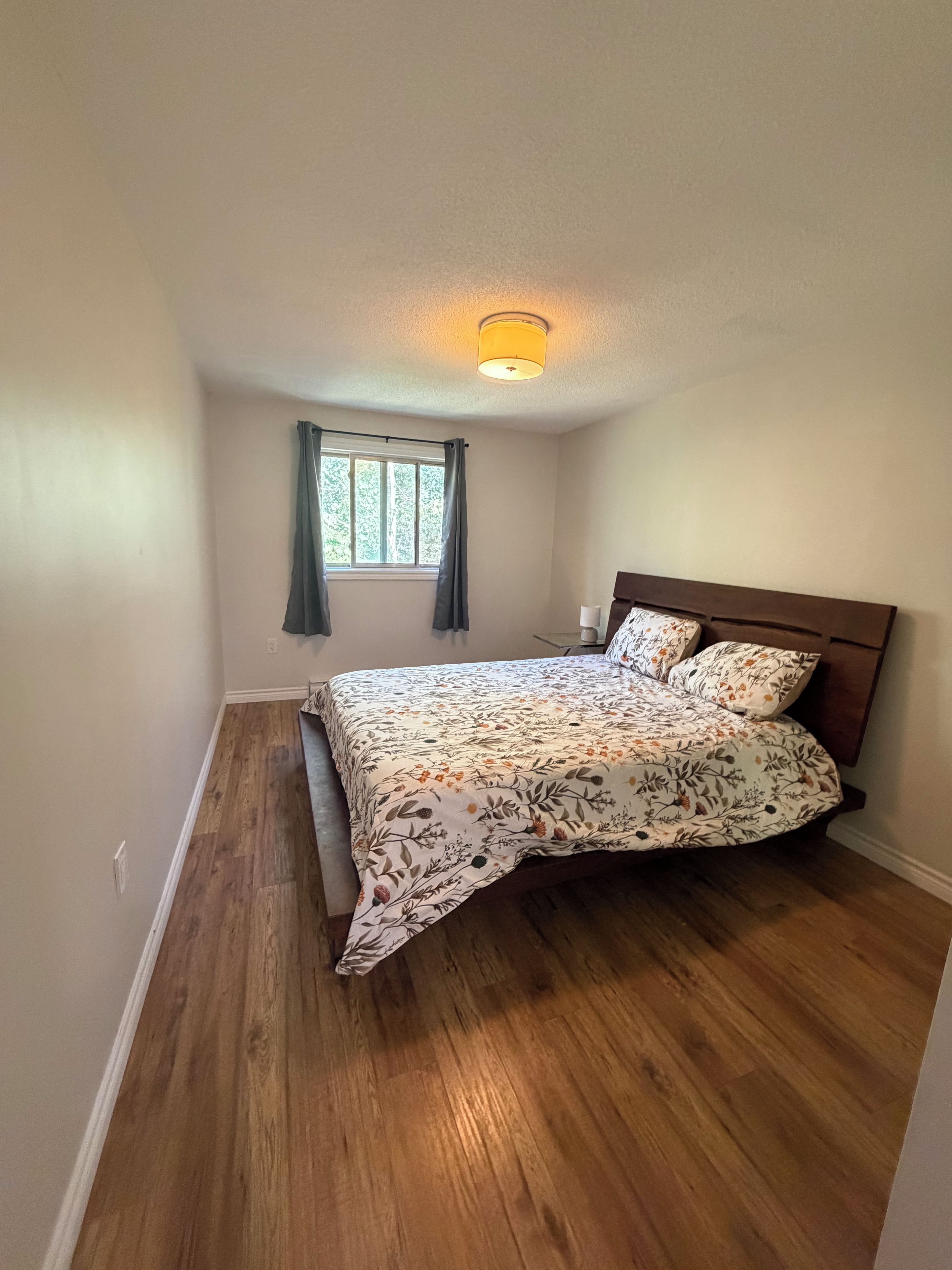 A bedroom with a wooden floor, a bed with floral bedding, a window with curtains, and a ceiling light.