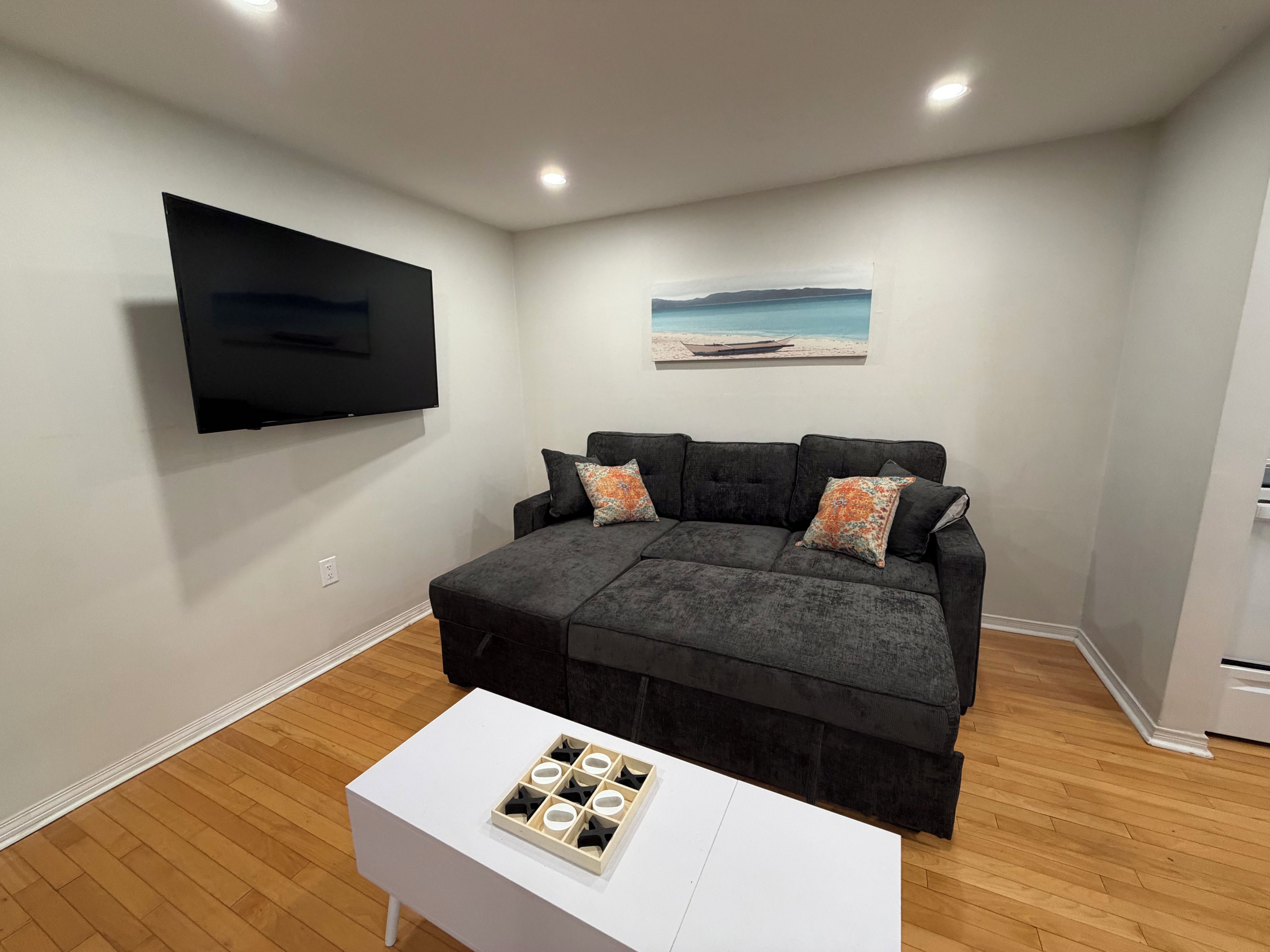 A living room with a grey sectional sofa, a flat-screen TV mounted on the wall, a small white table with a tray of candles, and a painting of a beach scene.