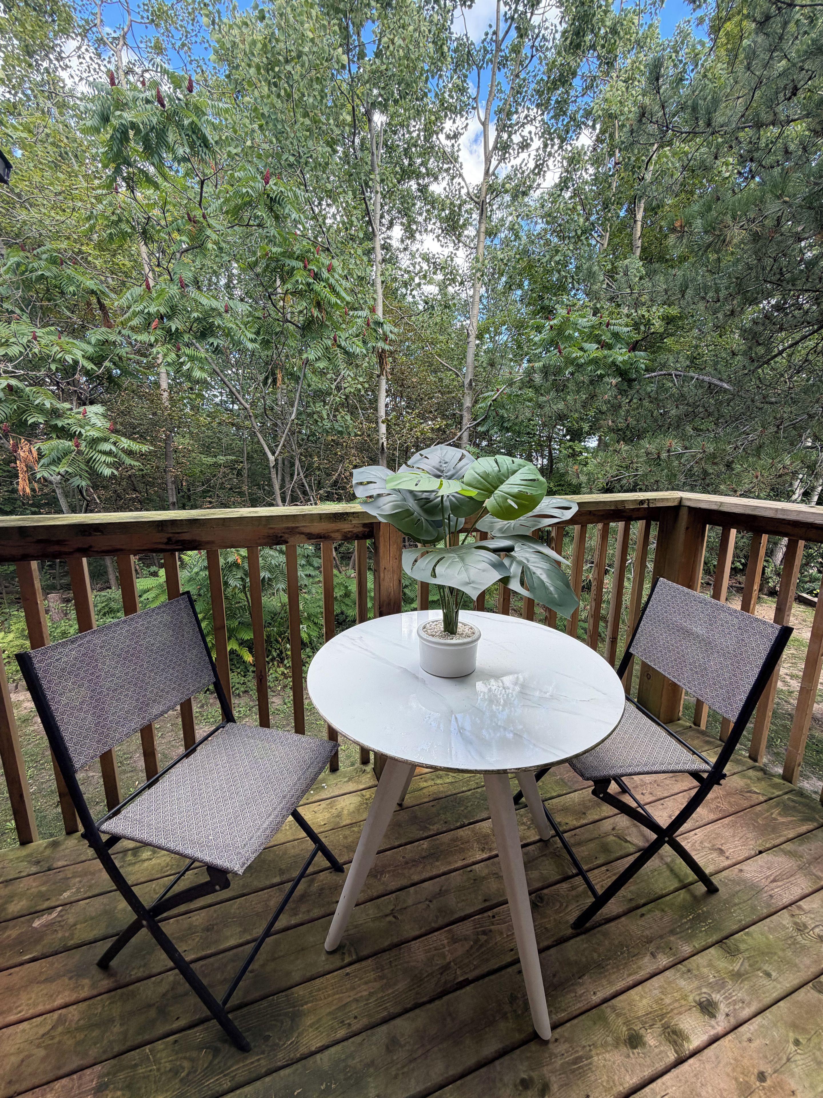 Una terraza de madera con una mesa redonda y dos sillas, rodeada de exuberante vegetación y árboles.