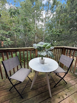 Una terraza de madera con una mesa redonda y dos sillas, rodeada de exuberante vegetación y árboles.