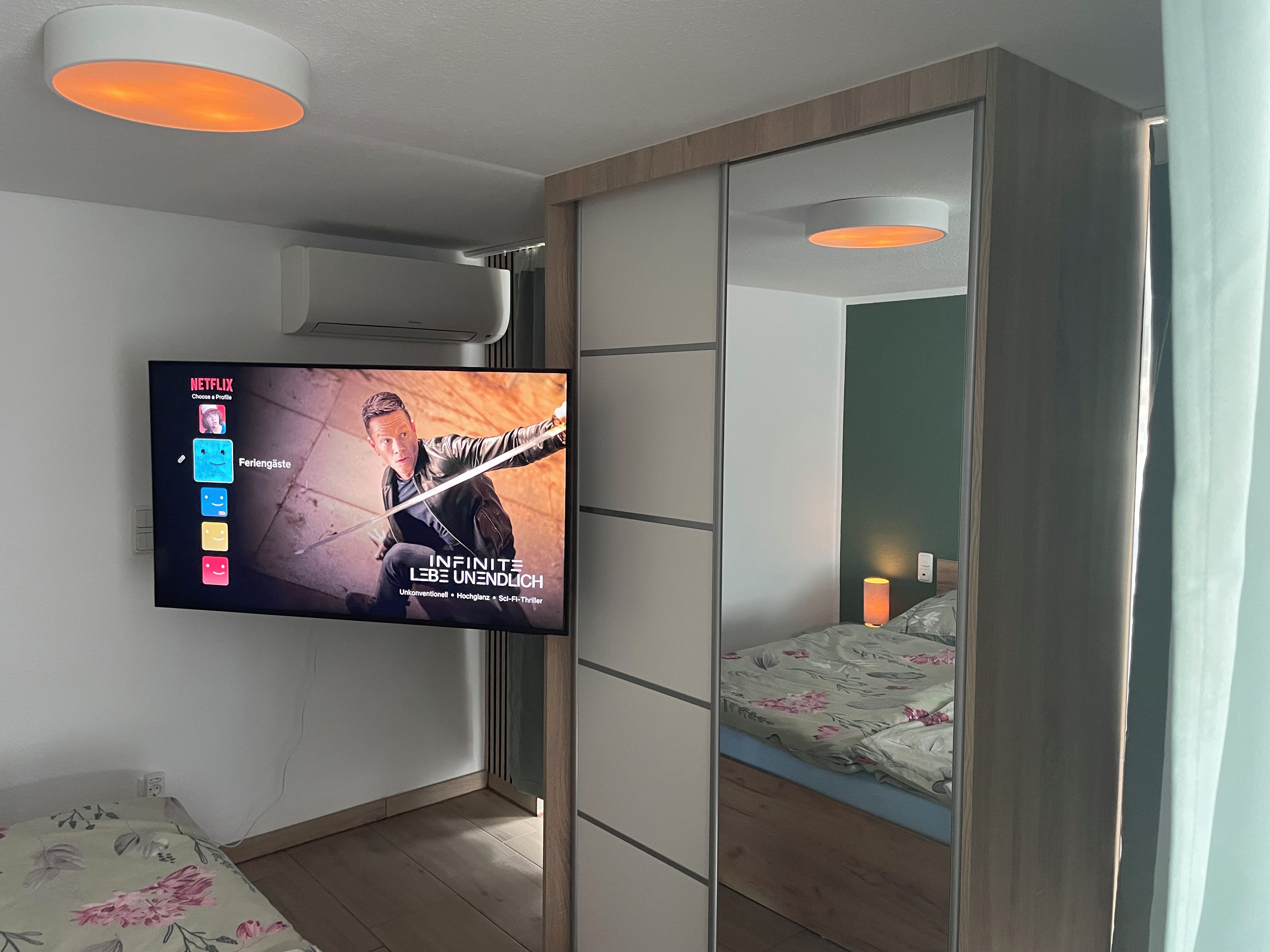 A modern bedroom with a TV mounted on the wall displaying a movie, a bed with floral bedding, and a bedside table.