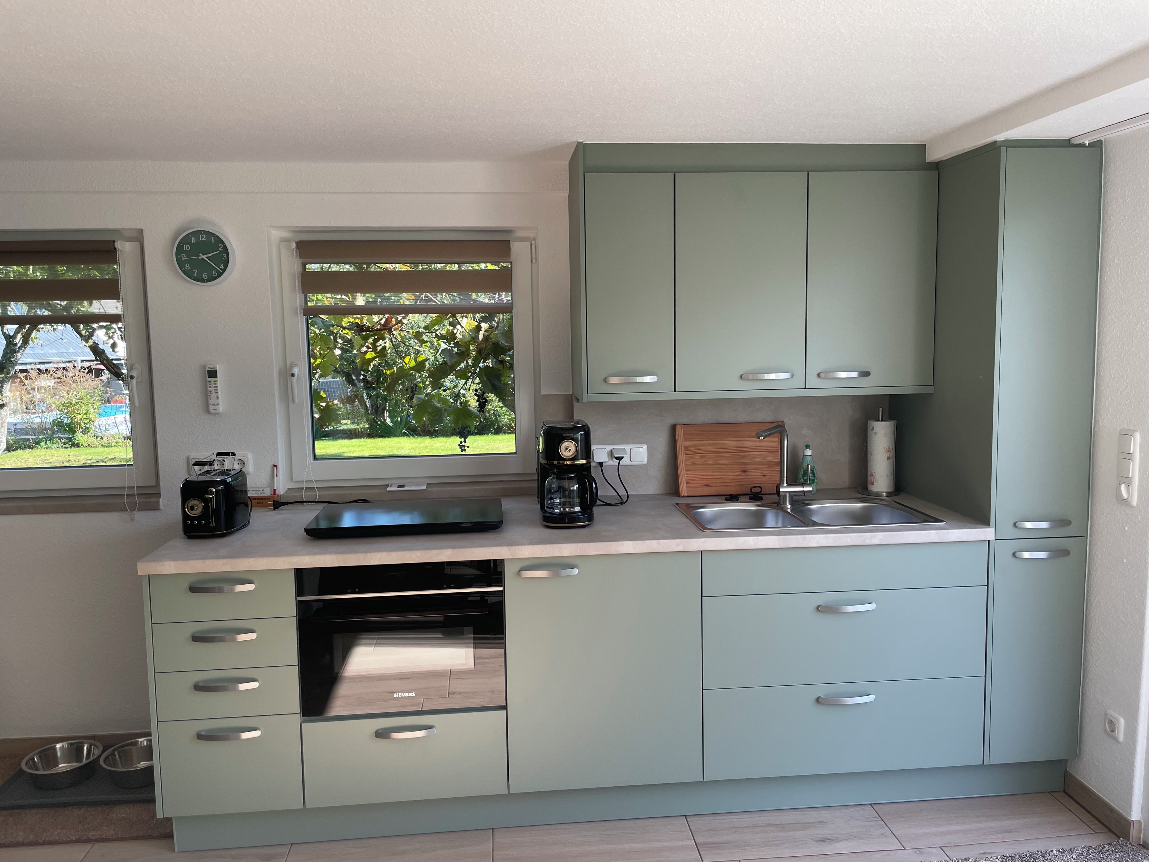 A modern kitchen with light green cabinets, a built-in oven, a coffee maker, and a window with a view of greenery.