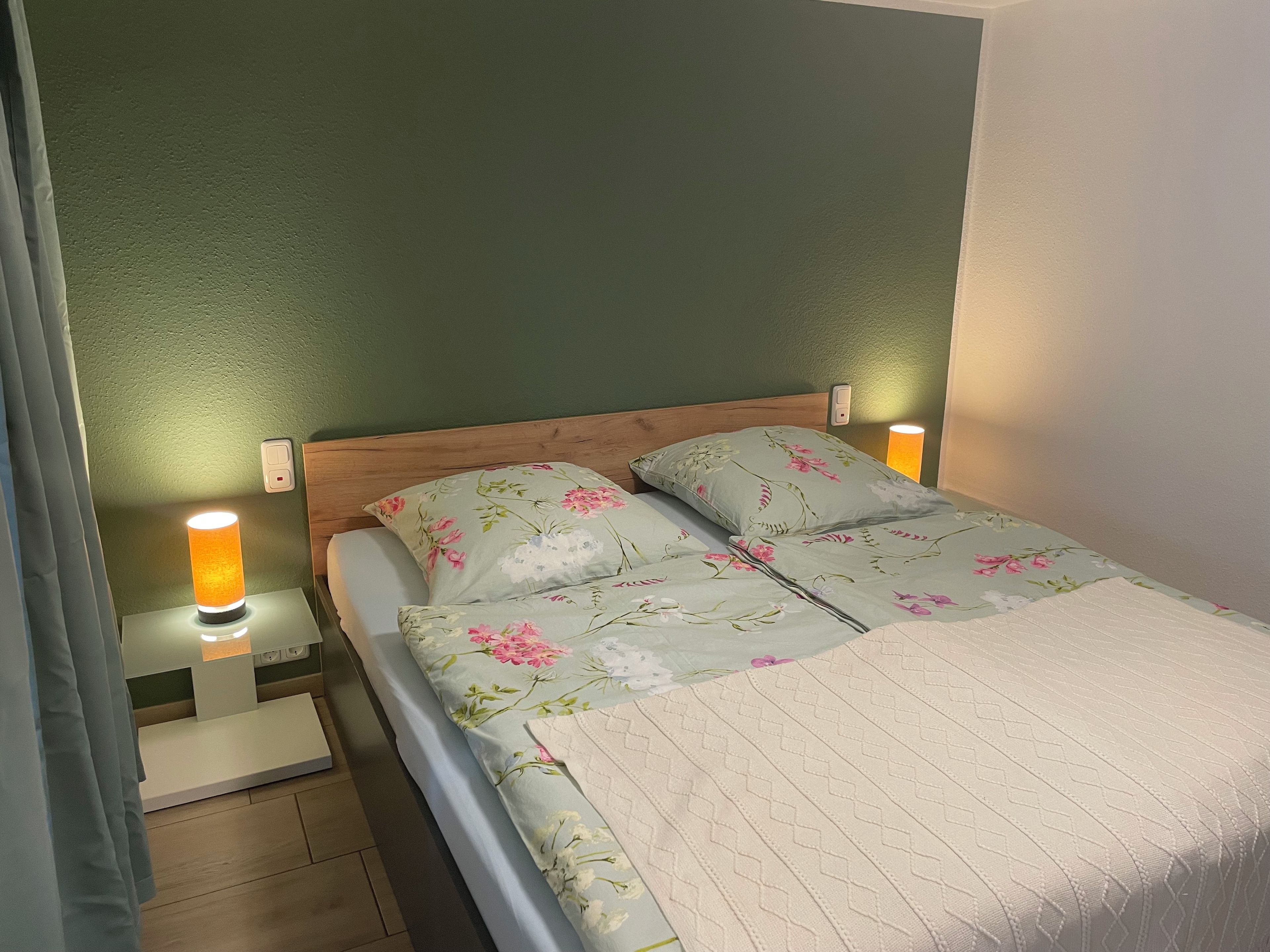 Ein Bett mit floralem Bettzeug, ein Holzkopfteil, Nachttischlampen und ein Nachttischchen mit Lampe.