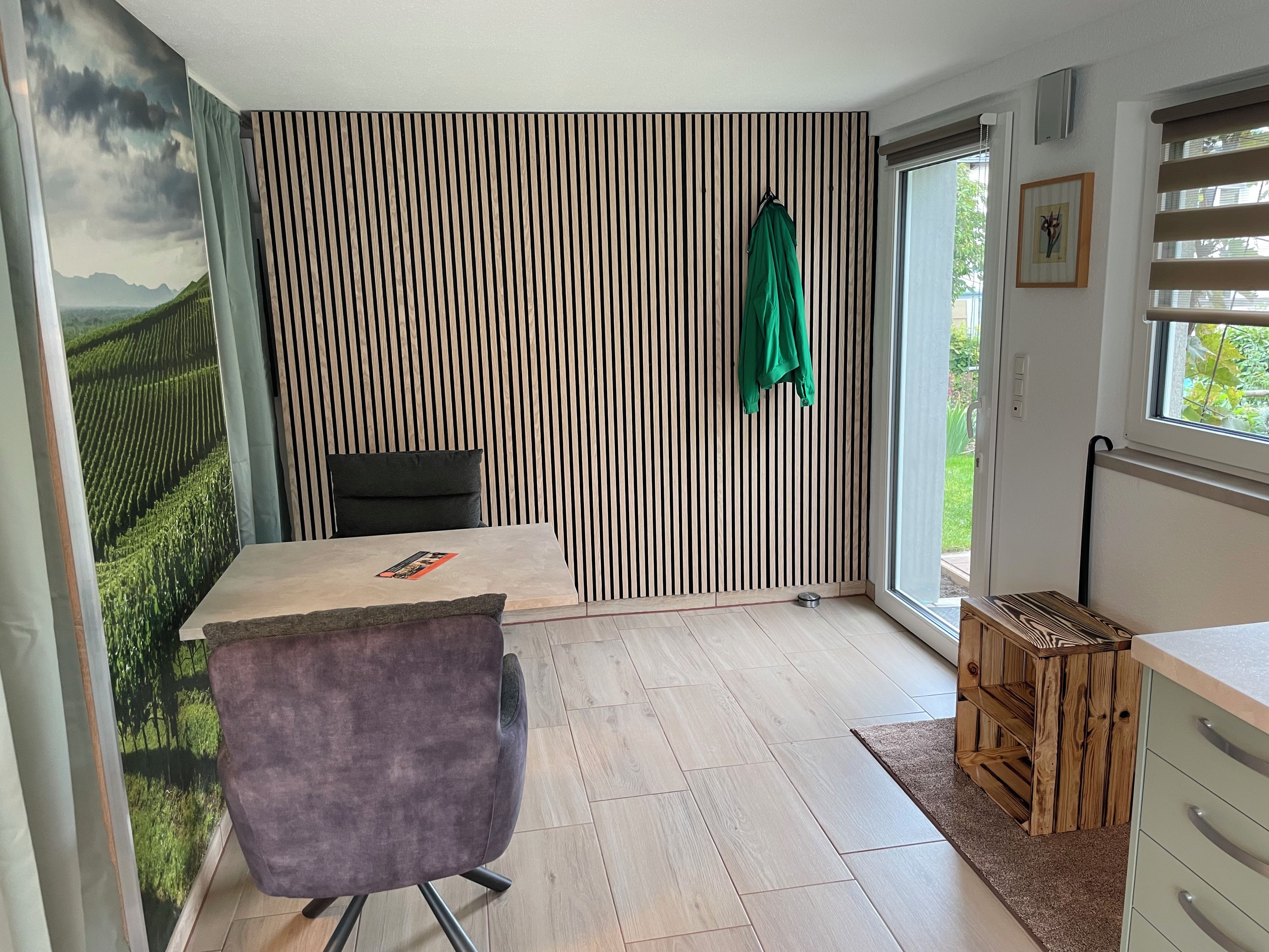 Ein Zimmer mit gestreifter Wand, einem Bett mit Kissen, einem Stuhl, einem kleinen Tisch, einem Fenster mit Jalousien und einer Tür.