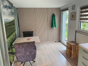 A room with a striped wall, a bed with a pillow, a chair, a small table, a window with blinds, and a door.