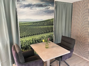 Ein Zimmer mit Tisch und zwei Stühlen, Blick auf einen Weinberg und einem Vorhang.