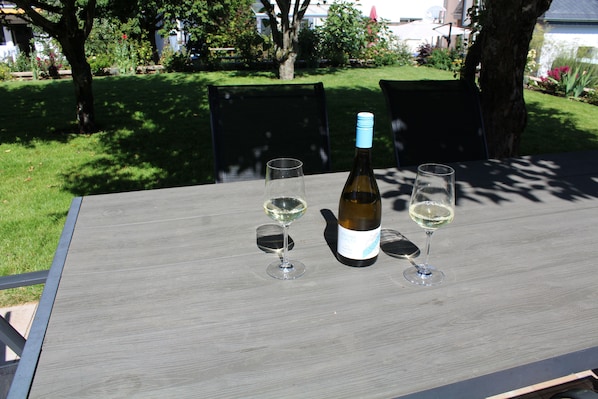 A table with a bottle and two glasses, set outdoors with a garden in the background.