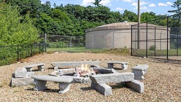 A fire pit with benches in a gravel area, surrounded by a fence and trees.