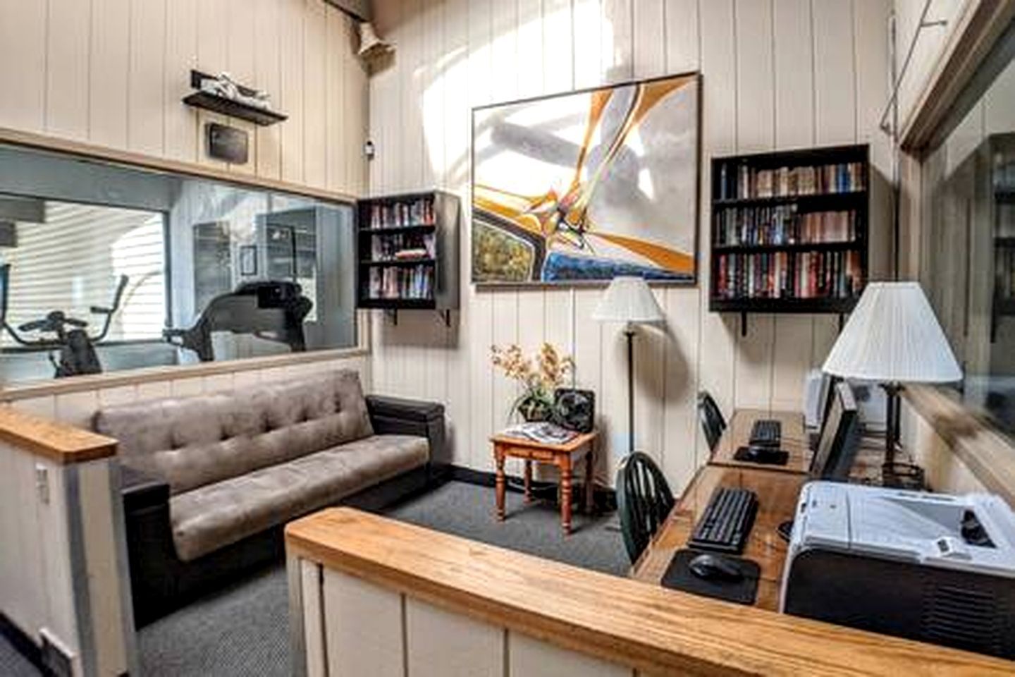 En kompakt opholdsstue med en sofa, et skrivebord med en computer, en bogreol og et stort vindue.