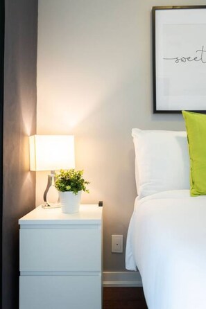 Ein ordentlich eingerichtetes Hotelzimmer mit Nachttisch, Lampe, Topfpflanze und einem Bett mit wei ßer Bettwäsche und einem grünen Kissen.