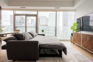 Quarto moderno com sofá-cama, televisão e vista para a cidade através de amplas janelas.