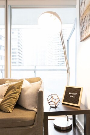 A modern living room with a sofa, a lamp, a welcome home sign, and a view of buildings through large windows. - Stylish 1BR APT near DWTN with Balcony Views (Toronto)