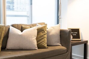 A sofa with multiple cushions, a framed welcome sign, and a window with blinds. - Stylish 1BR APT near DWTN with Balcony Views (Toronto)