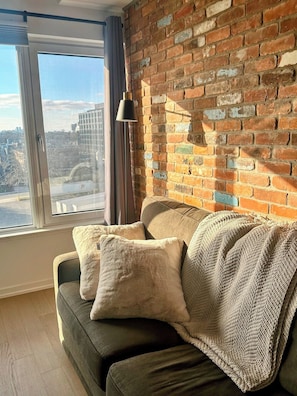 Une pièce avec un mur en briques, une fenêtre donnant sur un paysage urbain, un canapé avec des coussins et un lampadaire.