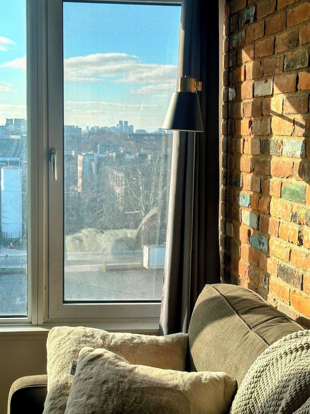 A room with a view of a cityscape through a window, a lamp, and a brick wall.