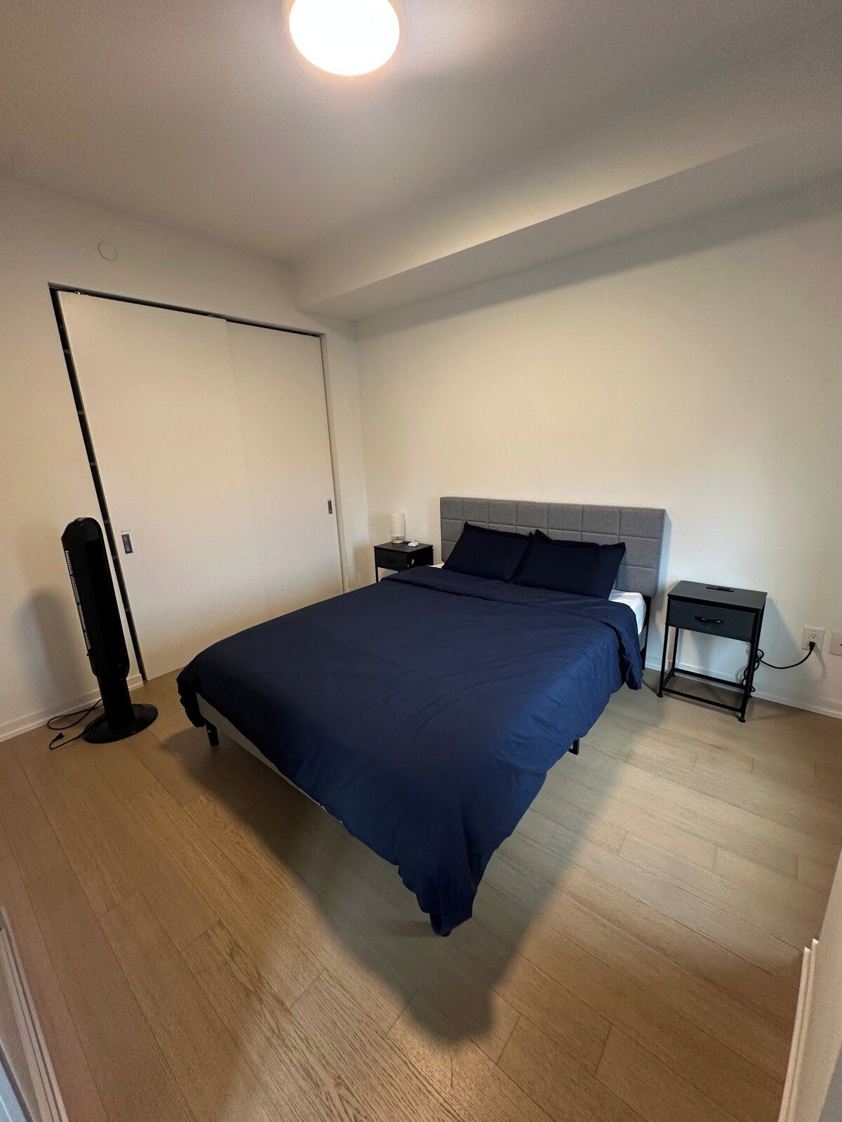Un dormitorio con cama, mesitas de noche, un ventilador y una puerta.