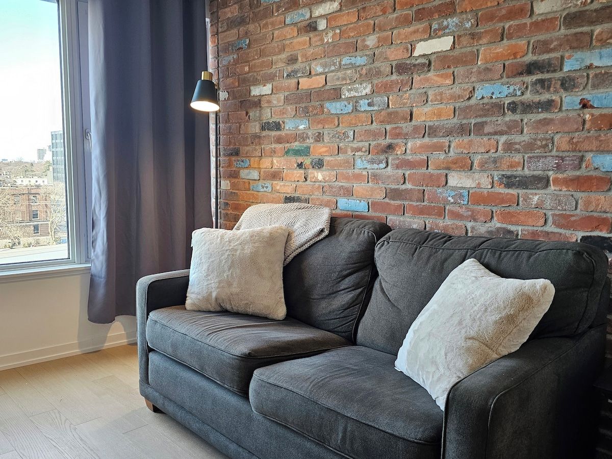 A grey sofa with two cushions against a brick wall, a window with curtains, and a pendant light.