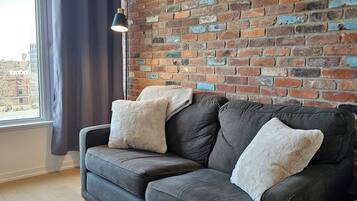 A grey sofa with two cushions against a brick wall, a window with curtains, and a pendant light.