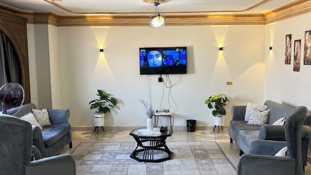 Een woonkamer met twee grijze banken, een glazen salontafel, een flatscreen televisie en decoratieve wandlampen.