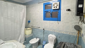 Een badkamer met een bad, twee toiletten, een wastafel, een douche en een raam met een ventilator.