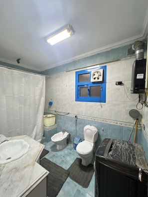 A bathroom with a bathtub, two toilets, a sink, a shower, and a window with a fan. - Degla Royal Residence : 2 BDR In Heart Of Degla (Cairo Governorate)