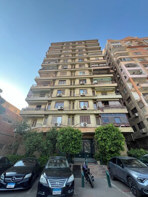 A multi-story residential building with balconies, air conditioning units, and a parking area with cars and a motorcycle. - Degla Royal Residence : 2 BDR In Heart Of Degla (Cairo Governorate)