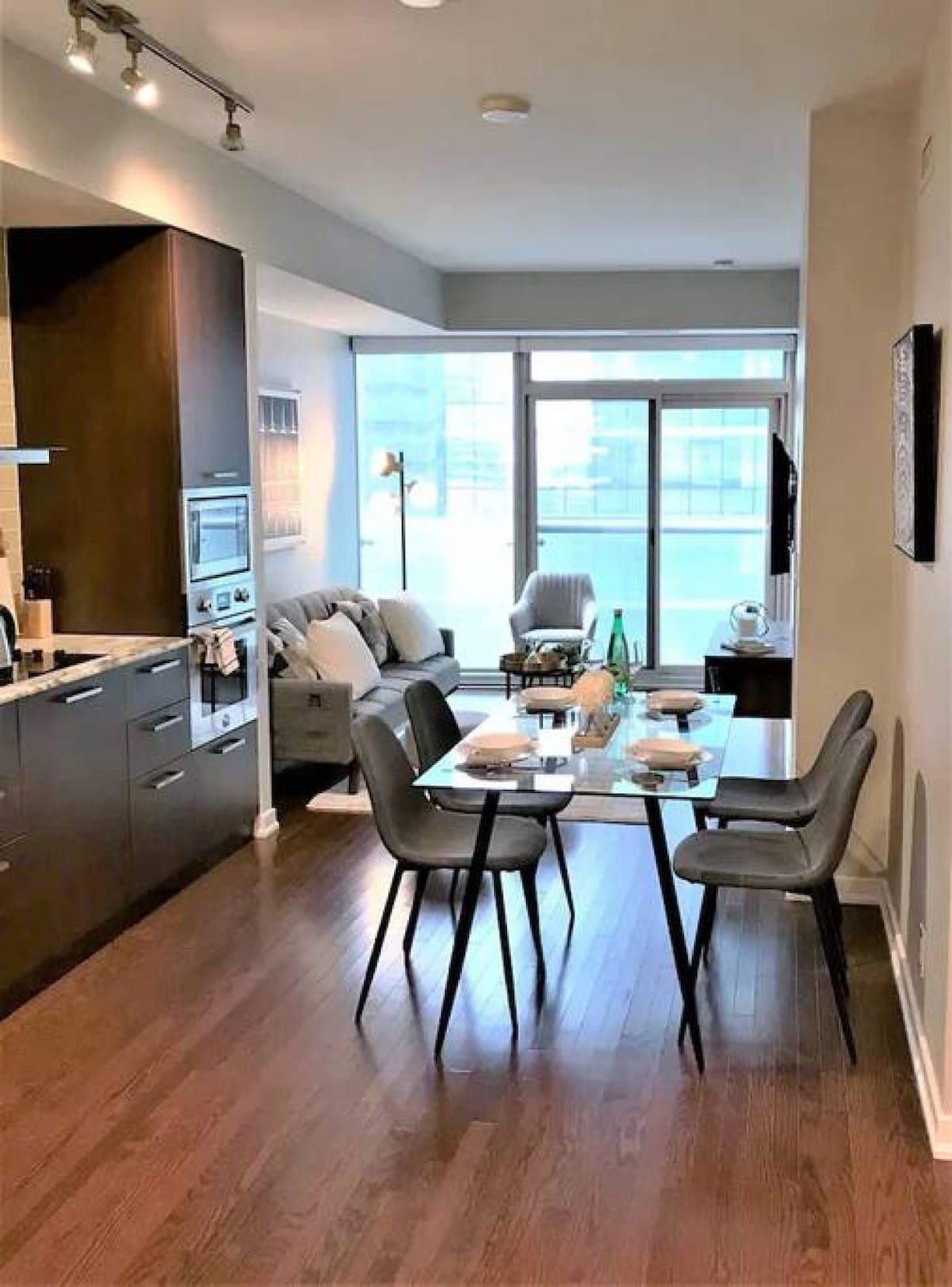 A modern kitchen with dark cabinetry, a dining area with a glass table, and a large window providing a view of the city.